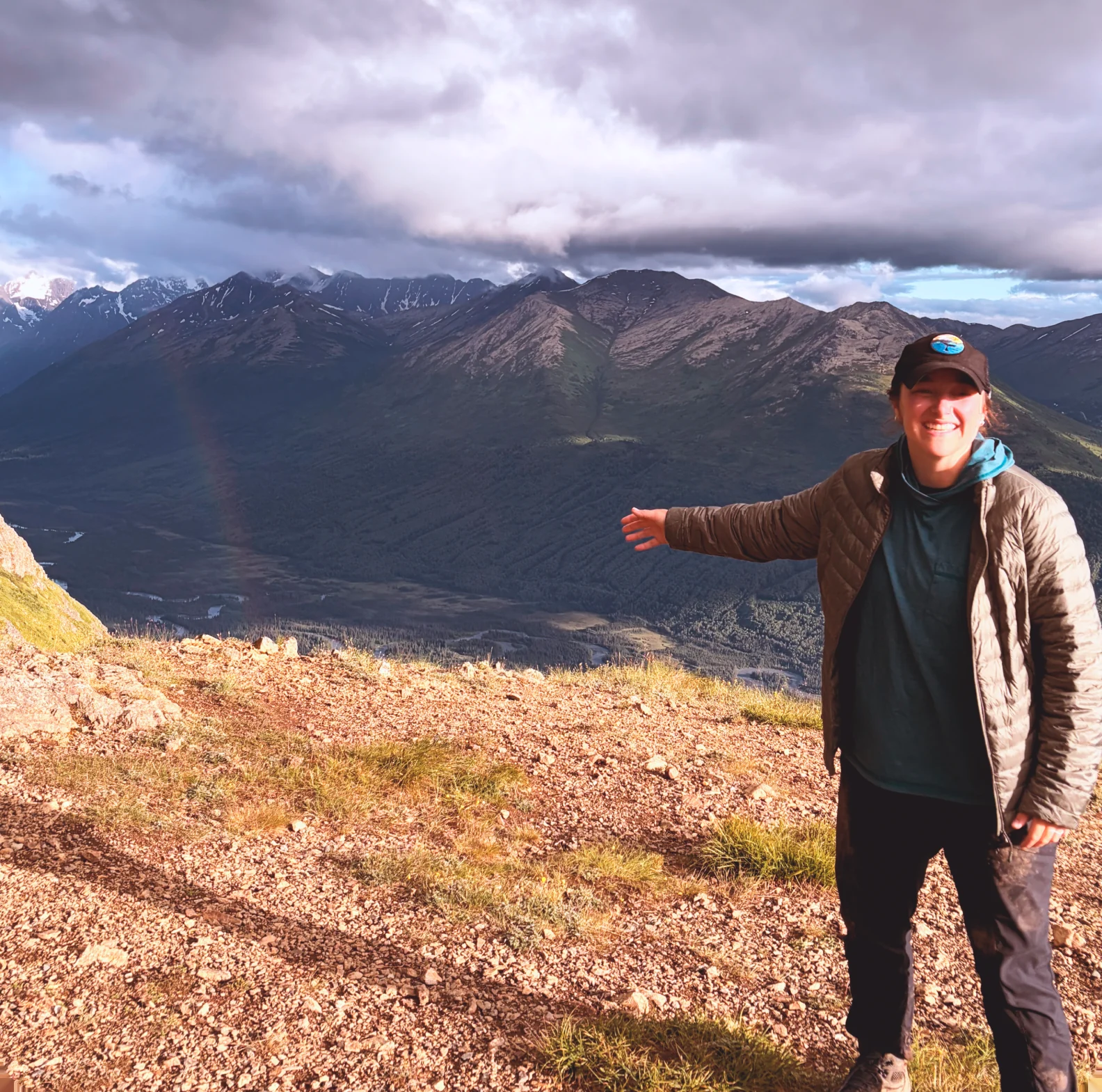 Morgan standing on an open tundra ridge with one arm outstretched and dark mountains behind