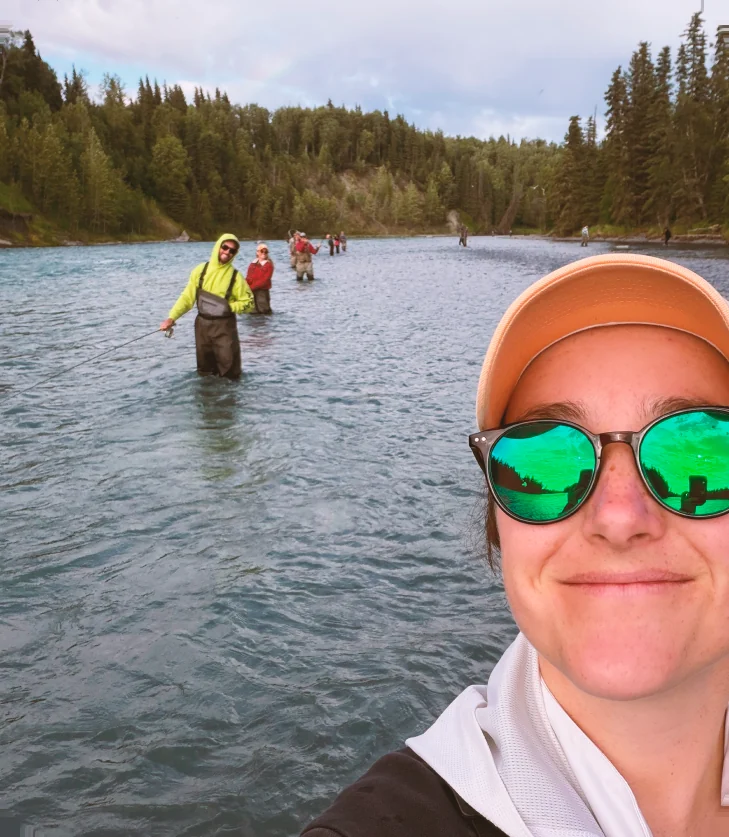 Close portrait of Morgan in a cap and mirrored sunglasses, with anglers wading the river behind