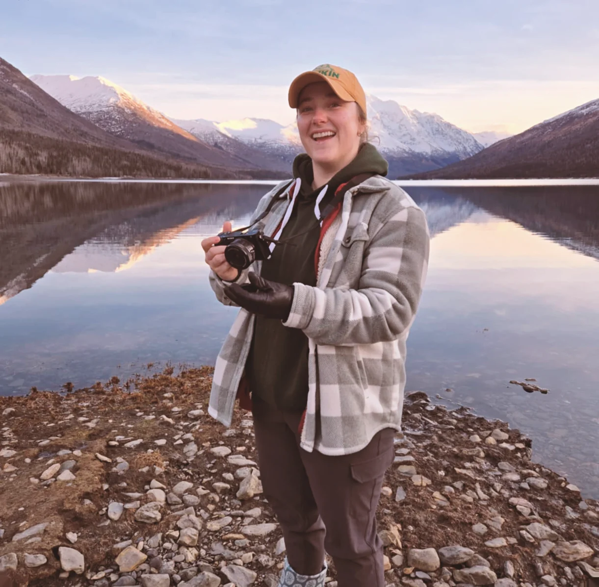 Morgan holding a camera on the gravel shore of a still lake at first light, mountains reflected in the water