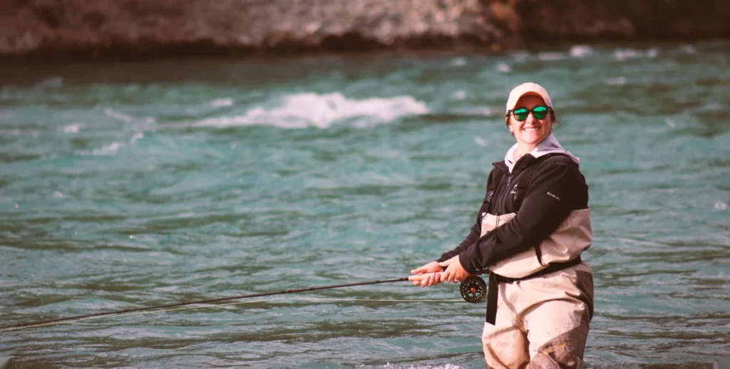 A person in waders casting a fly rod in a wide turquoise river beneath a dark rock face