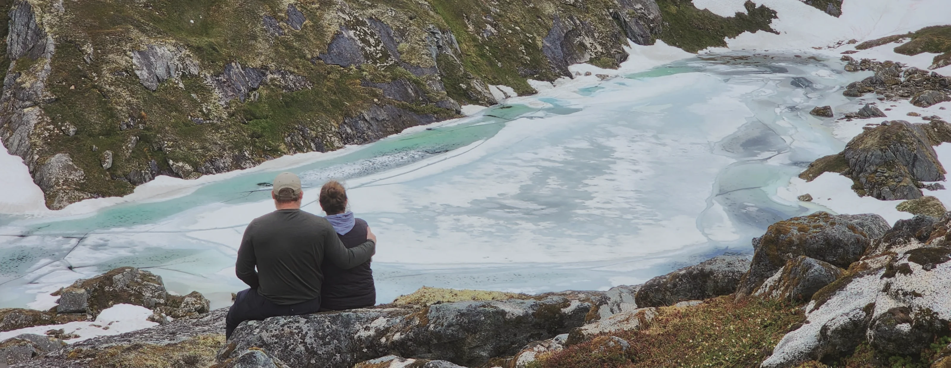 Two people sitting on rock looking down at a pale blue glacial lake ringed by scree